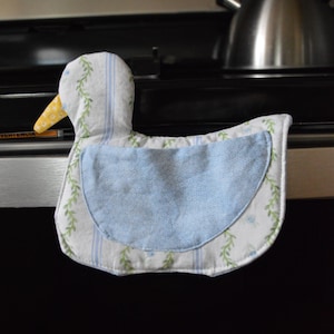 May include: A duck-shaped oven mitt. The mitt features a white background with blue stripes and green floral accents. The duck's bib is light blue with a small floral pattern. The beak is yellow. The mitt is approximately 8 inches long.
