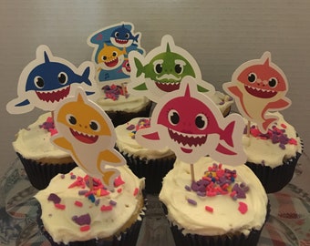 May include: Six cupcakes decorated with colorful sprinkles and topped with Baby Shark character cupcake picks. The cupcakes are on a white plate. A sign above the cupcakes reads "PARTY PICKS PLUS Handmade Cupcake Picks by Cassie and Jenny Omaha, NE 68104 Thank you for your business! Don't forget to leave a review."