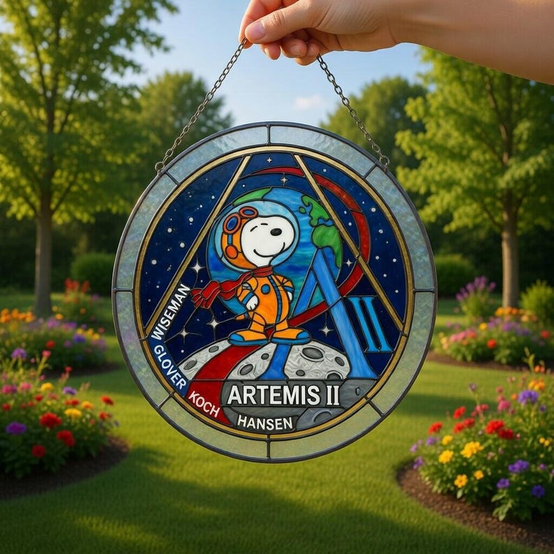 May include: A round stained glass window featuring Snoopy in an astronaut suit, standing on the moon with Earth in the background. The design includes the words "ARTEMIS II" and astronaut names. The window is held by a chain.
