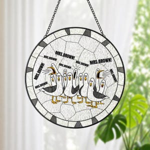 May include: A round, stained-glass window with a black metal frame and chain. The design features a flock of cartoon seagulls with the text "MRS. BROWN!" repeated around the edge. The seagulls are white with black wings and yellow feet.