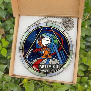 May include: A round stained glass window featuring Snoopy in an astronaut suit, with the text "ARTEMIS II". The window has a silver chain for hanging and is displayed in a cardboard box.