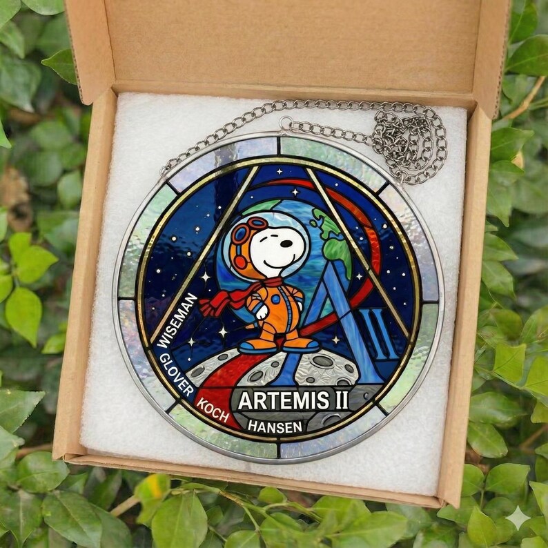 May include: A round stained glass window featuring Snoopy in an astronaut suit, with the text "ARTEMIS II". The window has a silver chain for hanging and is displayed in a cardboard box.