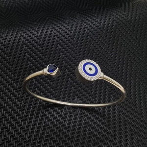 Può includere: Un braccialetto d'argento con un ciondolo blu di occhio turco e un ciondolo a forma di cuore blu. Il ciondolo di occhio turco è circondato da piccole pietre bianche.