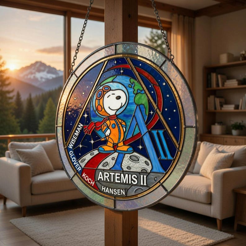 May include: A round stained glass window featuring Snoopy in an astronaut suit, with the text "ARTEMIS II". The design includes a blue background with stars, the Earth, and the names of the crew members. The window is hanging from a chain.