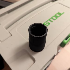 Peut inclure: Un adaptateur en plastique noir avec un intérieur texturé. L'adaptateur est cylindrique avec une base légèrement plus large, conçu pour les outils électriques. Il est posé sur une surface blanche avec le mot "TOOL" en vert.