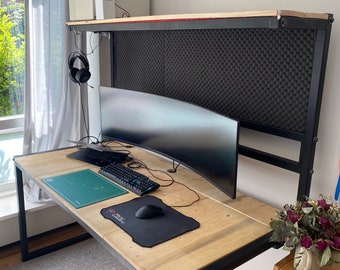 Può includere: Una scrivania con un telaio in metallo nero e un piano in legno. La scrivania ha un monitor curvo, una tastiera, un mouse e un tappetino da taglio verde. La scrivania è di fronte a un muro bianco con un pannello fonoassorbente nero dietro il monitor.