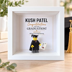 May include: A white shadow box frame containing a graduation-themed display. The background features the text KUSH PATEL Congratulations ON YOUR GRADUATION! UWE Bristol MEng Civil Engineering. A minifigure wearing a black graduation cap and gown holds a diploma.