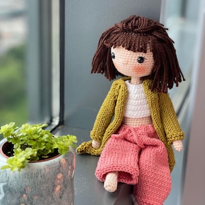 May include: A handmade crochet doll with brown hair and a mustard-colored cardigan. The doll wears a white top and pink pants. The doll is sitting on a windowsill next to a potted plant.