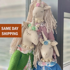 May include: Three handmade unicorn dolls with cream-colored bodies, each with a spiraled horn and closed eyes. They wear floral dresses and knitted sweaters in green, blue, and pink. Each doll has a name spelled out on a necklace.