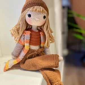 May include: A handmade crochet doll with blonde hair, wearing a brown beanie, a rust-coloured top, and brown trousers. The doll has a striped jacket with orange, yellow, and grey accents. The doll is sitting on a white surface.