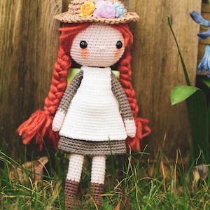 Op de afbeelding: Handgemaakte gehaakte pop met rood gevlochten haar, gekleed in een witte jurk, grijze rok en een strohoed versierd met kleurrijke bloemen. De pop staat in een grasveld bij een houten hek.