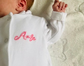 May include: A white baby onesie with pink embroidered lettering that says "Aaliyah".