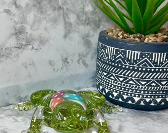 Könnte beinhalten: Eine durchscheinende grüne Froschfigur mit Glitzerakzenten und einem bunten Design auf dem Rücken. Der Frosch befindet sich vor einem dekorativen dunkelblauen Topf mit weißem geometrischen Muster und einer grünen Pflanze.