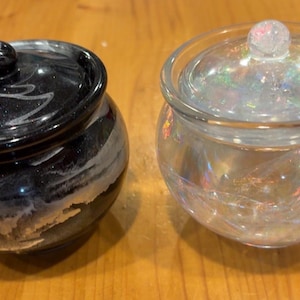 Könnte beinhalten: Zwei dekorative Gläser mit Deckel. Ein Glas ist schwarz mit weißer Marmorierung, das andere ist transparent mit schillerndem Schimmer. Beide Gläser haben eine runde Form und einen kleinen Knopf auf dem Deckel.