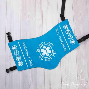 Puede incluir: Un chaleco azul brillante para perro de asistencia con correas y hebillas negras. El chaleco tiene texto blanco que dice "DO NOT PET" y "Assistance Dog". También presenta varios símbolos blancos.