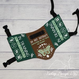 Puede incluir: Un arnés para perro de color verde oscuro y marrón con correas negras ajustables y hebillas de plástico. El texto dice IN TRAINING DO NOT DISTRACT y DO NOT DISTRACT distracting me can harm my handler, junto a iconos y una polilla.