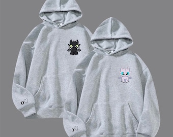 May include: Two gray hoodies with matching designs. Each hoodie features a cartoon dragon on the chest and initials with a heart on the sleeve. One dragon is black, the other is white and blue. Both have a hood and a front pocket.