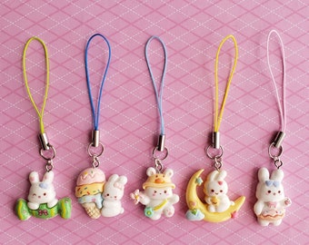 May include: Six colorful phone charms featuring cute bunny characters. Each charm has a different design, including a bunny holding a candy cane, a bunny with ice cream, a bunny with a flower, a bunny on a crescent moon, and two bunnies with different outfits.