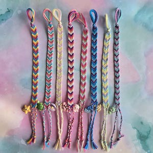 May include: Seven colorful friendship bracelets with chevron patterns and small charms. The bracelets are made with embroidery floss and have tassels at the ends.