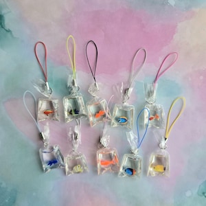 May include: A collection of miniature fish charms, each encased in a clear plastic bag, with colorful fish inside. Each bag has a loop with a different colored string, including red, yellow, black, and pink. The background is a pastel watercolor wash.