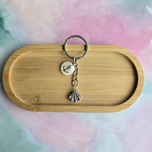 May include: A silver keychain with a round anchor charm and a seashell charm. The keychain is displayed on a wooden oval tray against a pastel watercolor background.