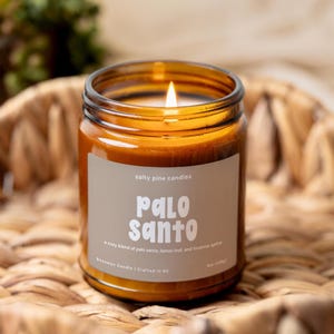 May include: A lit candle in an amber glass jar with a beige label reading Salty Pine Candles Palo Santo, a cozy blend of palo santo, lemon leaf, and incense spice, Beeswax Candle | Crafted in NC, 8oz (226g).