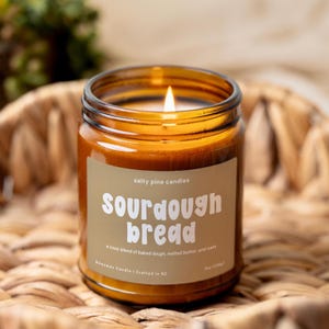 May include: A lit candle in an amber glass jar with a tan label. The label reads Salty Pine Candles, Sourdough Bread, a cozy blend of baked dough, melted butter, and oats, Beeswax Candle | Crafted in NC, 8oz (226g).