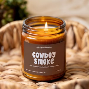 May include: A lit candle in an amber glass jar with a brown label reading Salty Pine Candles, Cowboy Smoke, a cozy blend of tobacco leaf, fireside embers, and rum, Beeswax Candle | Crafted in NC, 8oz (226g).