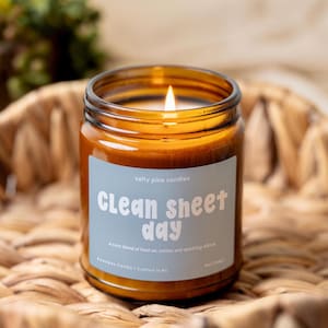May include: A lit candle in an amber glass jar with a light blue label. The label reads Salty Pine Candles, Clean sheet day, a cozy blend of fresh air, cotton, and sparkling citrus, Beeswax Candle | Crafted in NC, and 8oz (226g).