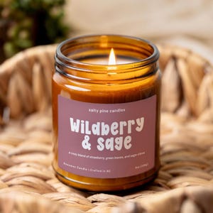 May include: A lit candle in an amber glass jar with a mauve label. The label reads Salty Pine Candles, Wildberry & Sage, a cozy blend of strawberry, green leaves, and sage clove, Beeswax Candle | Crafted in NC, and 8oz (226g).