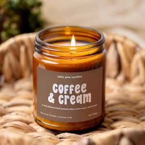 May include: A lit beeswax candle in an amber glass jar with a brown label. The label reads Salty Pine Candles, Coffee & Cream, a cozy blend of fresh brewed coffee, brown sugar, and cream, Beeswax Candle | Crafted in NC, and 8oz (226g).