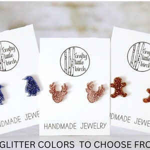 May include: Three pairs of glitter earrings on white cards. The earrings are shaped like penguins, reindeer, and gingerbread men. The cards have the text "crafty little birch" and "handmade jewelry".