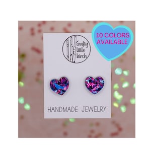 May include: A pair of heart-shaped earrings with a blue and pink iridescent finish. The earrings are handmade and come in 10 colors. The earrings are displayed on a white card with the text "crafty little birch" and "HANDMADE JEWELRY".