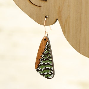 May include: A pair of wooden earrings with a green and brown painted pine tree design.