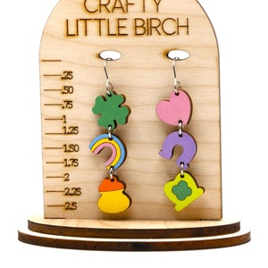 May include: Wooden earring display stand with the text "Crafty Little Birch." The stand features two pairs of dangling earrings with St. Patrick's Day themed charms, including a shamrock, rainbow, pot of gold, heart, horseshoe, and clover. The stand has a measurement scale.