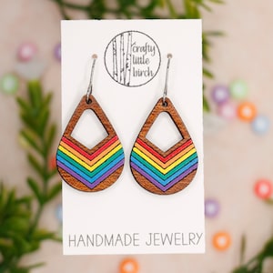 May include: Wooden teardrop earrings with a rainbow chevron design. The earrings are made of wood and feature a colorful, geometric pattern in red, orange, yellow, green, blue, and purple. The earrings hang from silver-colored hooks. The card reads "Handmade Jewelry".