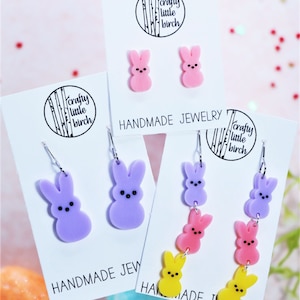May include: Three pairs of handmade bunny-shaped earrings. The first pair is pink, the second pair is purple, and the third pair is a stack of yellow, pink, and purple bunnies.