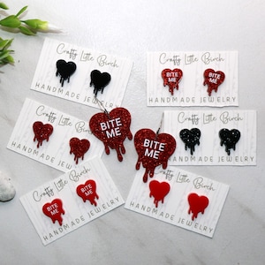 May include: Four pairs of red and black glitter heart-shaped earrings with the text "Bite Me" on the red hearts. The earrings are displayed on white cards with the text "Crafty Little Birch Handmade Jewelry".