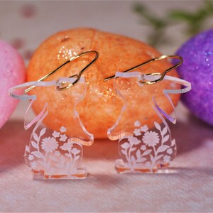 K&ouml;nnte beinhalten: Ein Paar transparente Acryl-Ohrringe in Form von Hasen mit Blumenmustern. Die Ohrringe haben goldene Haken.