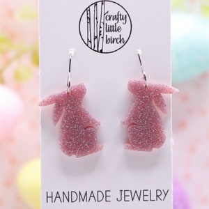 K&ouml;nnte beinhalten: Ein Paar rosa Glitzer-Hasenohrringe mit silbernen Kreolen. Die Ohrringe befinden sich auf einer wei&szlig;en Karte mit dem Text "crafty little birch" und "HANDMADE JEWELRY".