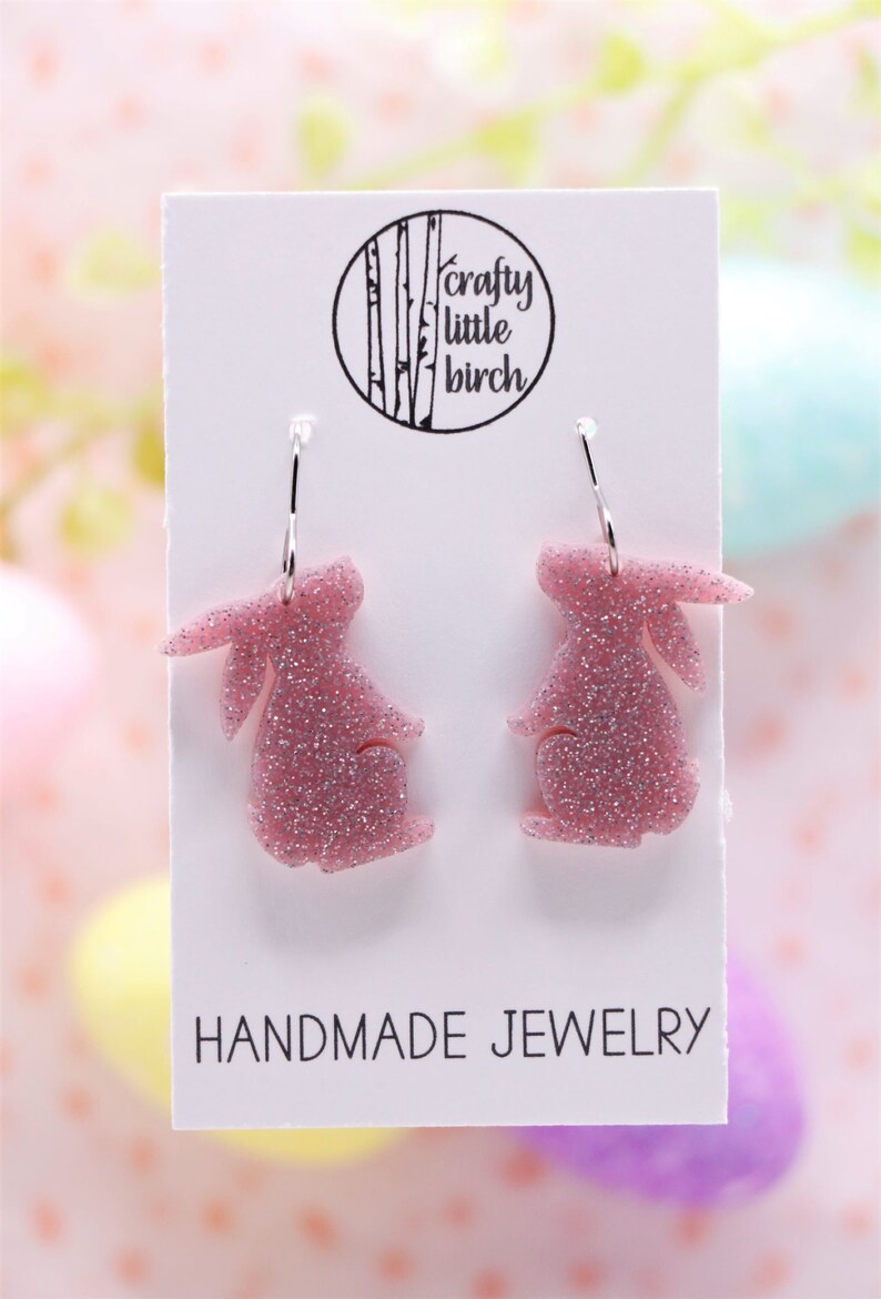 K&ouml;nnte beinhalten: Ein Paar rosa Glitzer-Hasenohrringe mit silbernen Kreolen. Die Ohrringe befinden sich auf einer wei&szlig;en Karte mit dem Text "crafty little birch" und "HANDMADE JEWELRY".