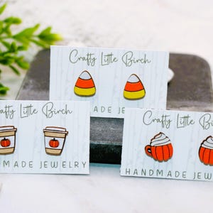 May include: Three sets of earrings on white cards with the text "Crafty Little Birch" and "Handmade Jewelry". The earrings are made of wood and feature fall-themed designs: a pumpkin spice latte, a candy corn, and a pumpkin.