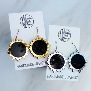 May include: Two pairs of sun-shaped dangle earrings on white display cards labeled Crafty Little Birch Handmade Jewelry. One pair features a gold-tone finish with black centers, while the other pair has a silver-tone finish with black centers.