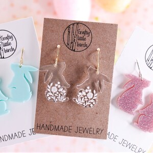 K&ouml;nnte beinhalten: Drei Paar handgefertigte Hasenohrringe. Das erste Paar ist hellblau, das zweite Paar ist klar mit wei&szlig;en Blumendesigns und das dritte Paar ist pink mit Glitzer. Jedes Paar befindet sich auf einer Karte mit dem Text "crafty little birch" und "handgemachter Schmuck".