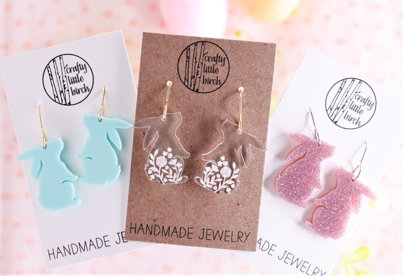 K&ouml;nnte beinhalten: Drei Paar handgefertigte Hasenohrringe. Das erste Paar ist hellblau, das zweite Paar ist klar mit wei&szlig;en Blumendesigns und das dritte Paar ist pink mit Glitzer. Jedes Paar befindet sich auf einer Karte mit dem Text "crafty little birch" und "handgemachter Schmuck".