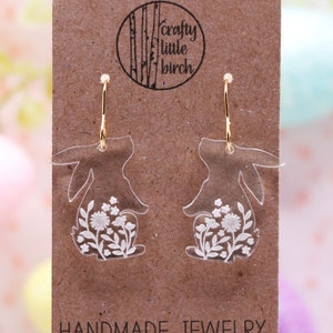 K&ouml;nnte beinhalten: Ein Paar transparente Acryl-Hasenohrringe mit wei&szlig;en Blumendetails. Die Ohrringe befinden sich auf einer braunen Karte mit dem Text "crafty little birch" und "HANDMADE JEWELRY".