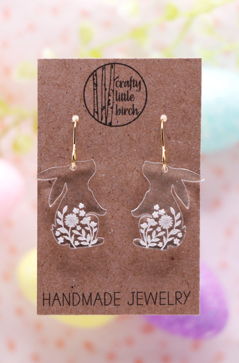 K&ouml;nnte beinhalten: Ein Paar transparente Acryl-Hasenohrringe mit wei&szlig;en Blumendetails. Die Ohrringe befinden sich auf einer braunen Karte mit dem Text "crafty little birch" und "HANDMADE JEWELRY".