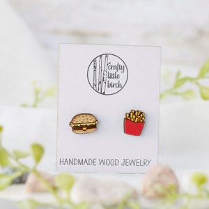 May include: A pair of wooden earrings featuring a burger and french fries. The earrings are handmade and are on a white card with the text "crafty little birch" and "HANDMADE WOOD JEWELRY".