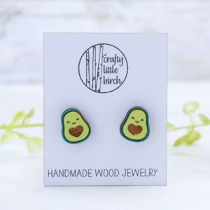 May include: A pair of wooden avocado earrings with a heart and a smiley face. The earrings are green, brown, and teal. The earrings are on a white card with the text "crafty little birch" and "HANDMADE WOOD JEWELRY".