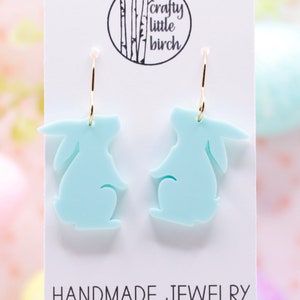 K&ouml;nnte beinhalten: Ein Paar hellblaue Ohrringe in Hasenform mit goldenen Kreisen. Die Ohrringe befinden sich auf einer wei&szlig;en Karte mit dem Text "crafty little birch" und "HANDMADE JEWELRY".